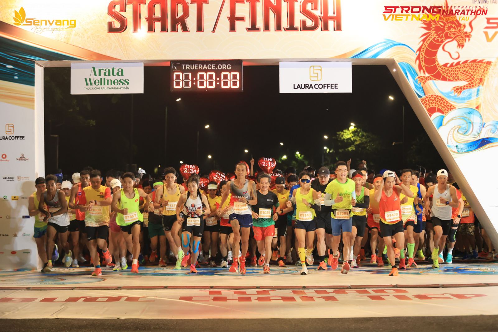 Runners surging through the start gate at night under the TrueRace clock